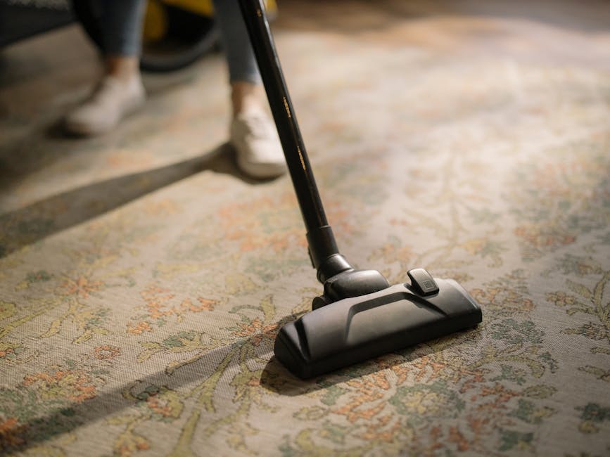 Close-up of a vacuum cleaner head being used to clean an intricate, multicolored patterned carpet in a room with natural lighting. The vacuum cleaner is black with a visible control button on top, and