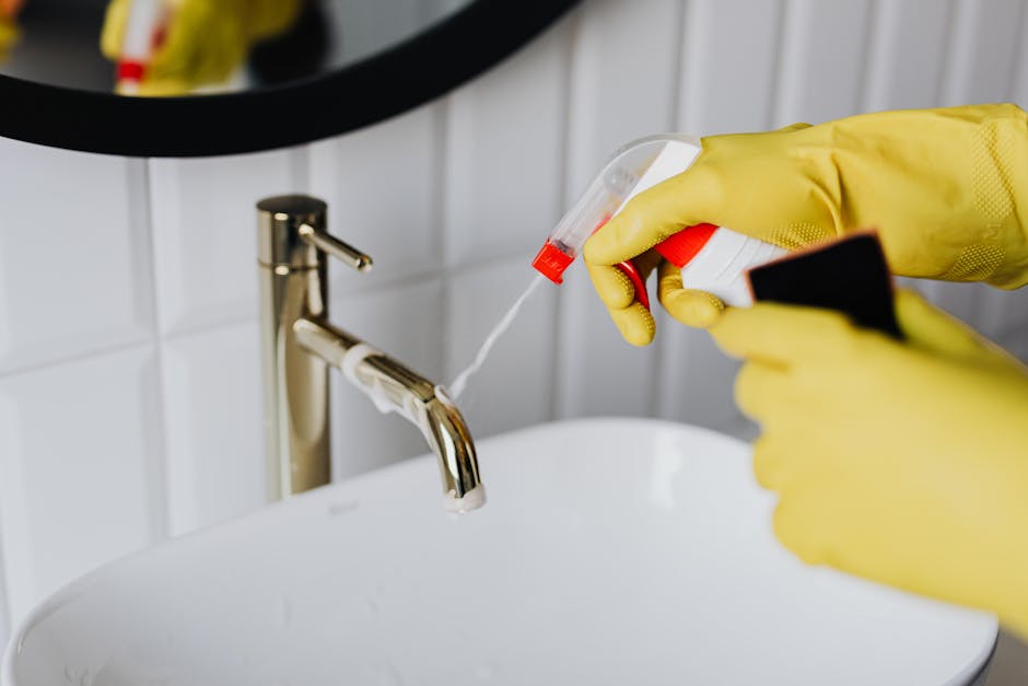 Close-up of a person's gloved hands holding a spray bottle filled with cleaning solution and a cloth, preparing to clean a white ceramic sink with a chrome faucet in a residential bathroom. The backgr
