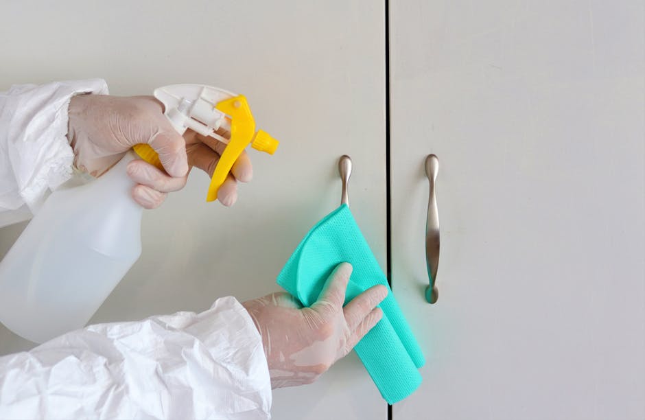 Close-up of a person's gloved hands holding a spray bottle filled with cleaning solution and a cloth, preparing to clean a white ceramic sink with a chrome faucet in a residential bathroom. The backgr