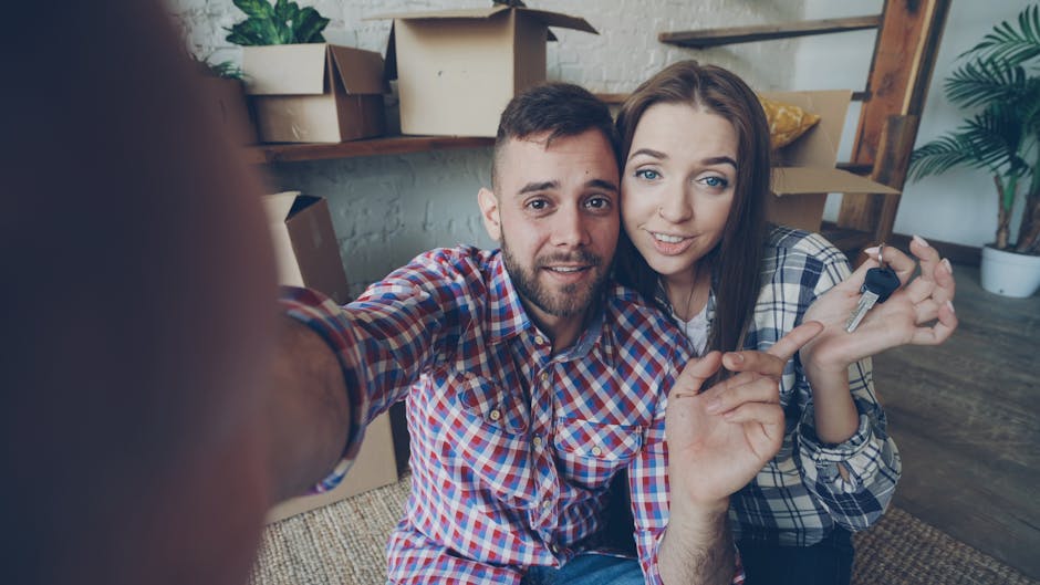 A young couple indoors, sitting on a wooden floor surrounded by cardboard moving boxes and potted plants, smiling and looking at the camera as they hold a set of keys, symbolizing moving out or handover. The room is well-lit with natural light, with visible surfaces including a textured white brick wall and a wooden shelf in the background. Both individuals are casually dressed, and the scene suggests a clean and tidy environment, aligning with the context of end of tenancy cleaning. As part of professional cleaning services, Cleaners Kingston specialises in thorough surface cleaning and hygiene for moving purposes, ensuring properties are spotless for new occupants.