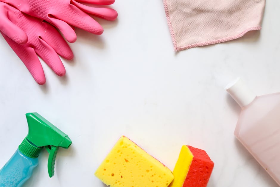 A flat lay of cleaning tools and supplies for surface cleaning and sanitisation, including a pair of pink rubber gloves, a green and blue spray bottle, a yellow and red sponge, a pink microfiber cloth, and a white cleaning spray bottle, arranged on a white surface. The items are neatly positioned, with the gloves at the top left, the sponge at the bottom center, and the cleaning bottles and cloth around them, illustrating a professional domestic cleaning setup. The lighting is bright and even, emphasizing the cleanliness and readiness for thorough cleaning tasks, aligning with content on end of tenancy cleaning in Kingston, provided by Cleaners Kingston.