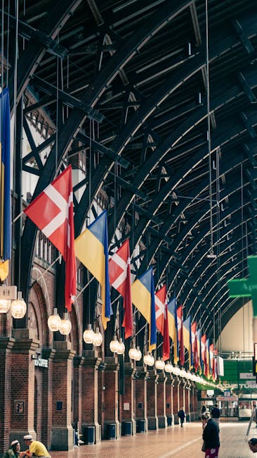 Interior view of Kingston Station featuring a high, arched ceiling with dark metal beams and structural supports. Several multicolored flags, including red, yellow, white, and blue, are hanging from the ceiling along the length of the station. The brick wall beneath the flags has a series of evenly spaced, glowing spherical lights attached to metal fixtures. The station floor is smooth and clean, with a few passengers walking or waiting, contributing to a tidy and well-maintained atmosphere. Cleaners Kingston provides surface and deep cleaning services to maintain the hygiene and appearance of such public spaces, ensuring a safe and welcoming environment.