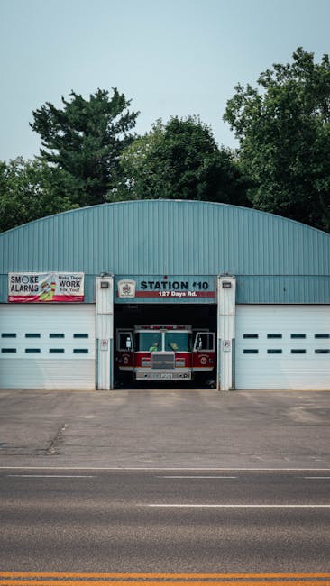 A fire station with a rounded, blue-colored metal exterior and a large garage door in the center, which is open revealing a red fire truck inside. The fire truck is clean and well-maintained, featuring a silver grille and emergency lights on top. Above the garage, there's a sign marked 'STATION 10' and street address '127 Daya Rd.' Against the green treetops in the background, the station appears bright and orderly, with no visible dirt or clutter on the building or pavement. The scene is lit with natural daylight, emphasizing the tidy and professional appearance of the station, reflecting the importance of hygiene and maintenance which are key aspects of residential and commercial cleaning services provided by Cleaners Kingston.